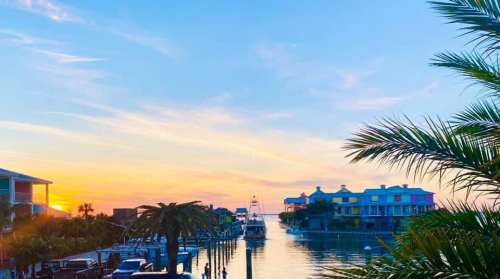 A serene waterfront scene at sunset, featuring colorful houses, palm trees, and calm waters reflecting the sky.