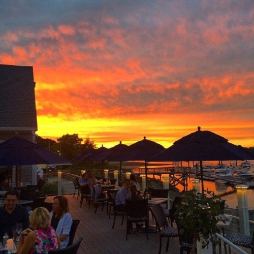 A vibrant sunset over a marina, with diners under blue umbrellas enjoying the view.