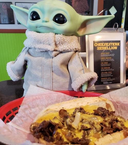 A toy figure resembling a green alien stands next to a cheesesteak sandwich with melted cheese on a red tray.