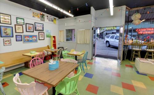 Colorful cafe interior with tables, chairs, and framed artwork, featuring an open door leading outside.