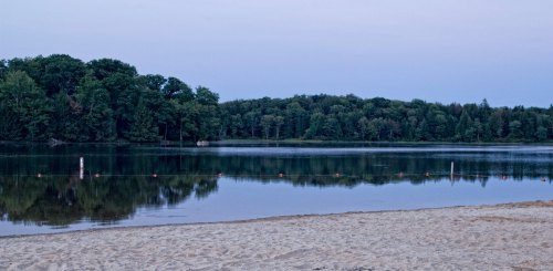 A serene lake at dusk, surrounded by trees, with a sandy beach in the foreground and calm water reflecting the landscape.