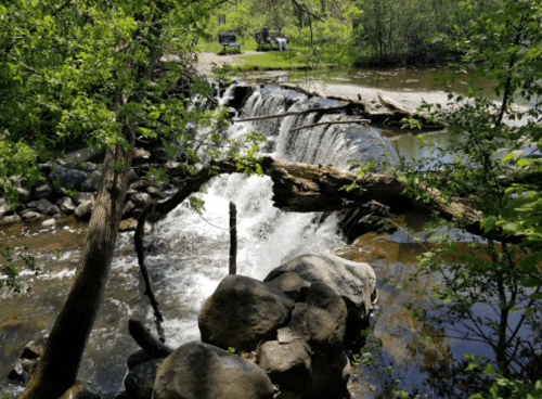 A serene waterfall cascades over rocks, surrounded by lush greenery and a peaceful park with benches in the background.