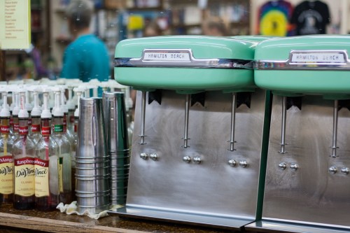Vintage Hamilton Beach drink mixers in mint green, alongside bottles of syrups and metal cups on a wooden counter.