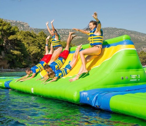 A group of five people in life jackets jumping off a colorful inflatable water slide into a clear blue lake.