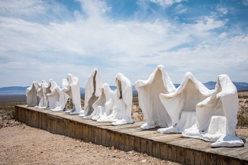A series of white, ghostly sculptures on a wooden platform in a desert landscape under a blue sky.