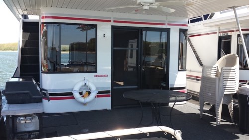 A houseboat with a deck, table, and chairs, overlooking a calm body of water on a sunny day.