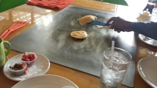 A hand flips a pancake on a griddle, surrounded by plates of toppings and a glass of water on a wooden table.