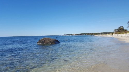 A serene beach scene with clear water, a large rock, and a distant shoreline under a bright blue sky.