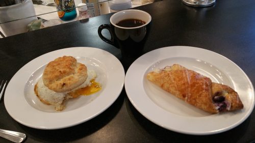 A black coffee cup beside a plate with a biscuit and egg, and another plate with a blueberry pastry.