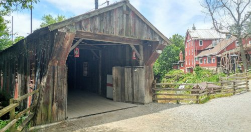 A rustic wooden covered bridge with an open entrance, surrounded by greenery and a red barn in the background.