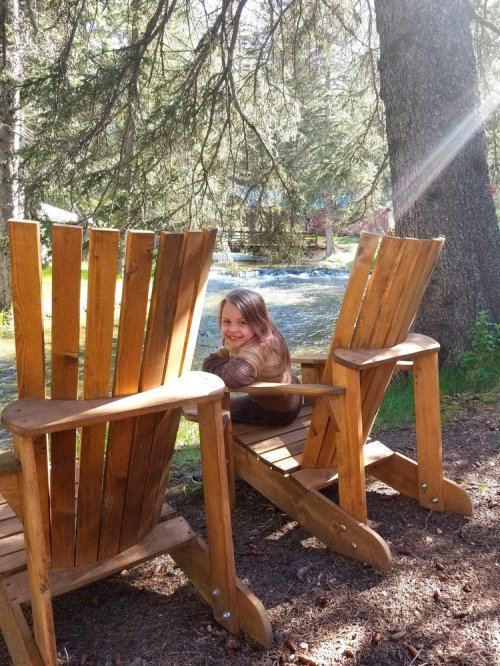 A girl smiles while sitting between two wooden chairs by a river, surrounded by trees and sunlight.
