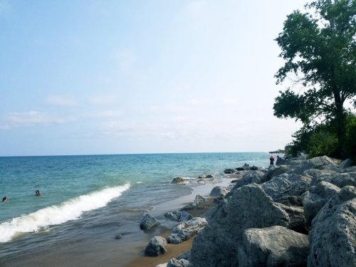 A serene beach scene with gentle waves, rocky shore, and a few people enjoying the water under a clear blue sky.