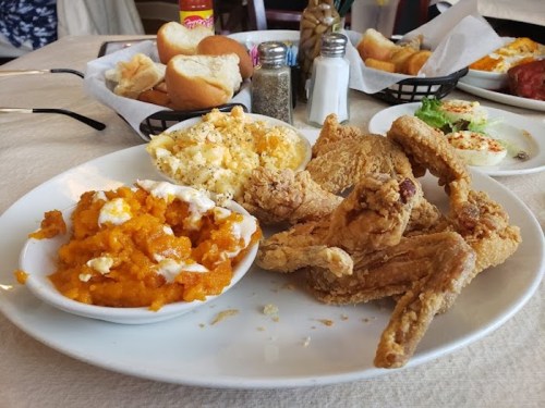 A plate of fried chicken with sweet potatoes and macaroni and cheese, accompanied by rolls and condiments in the background.