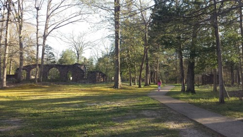 A girl walks along a path in a wooded area, with stone ruins and trees surrounding her in the background.