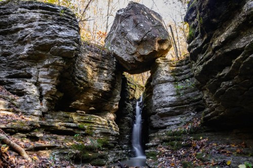 A serene waterfall flows between rocky cliffs, with a large boulder perched above in a forested setting.