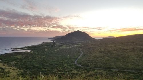 Sunset over a coastal landscape with rolling hills, a winding road, and a calm ocean in the background.