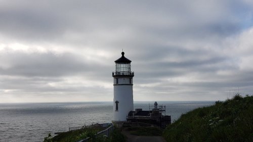 A white lighthouse stands on a grassy cliff overlooking a calm sea under a cloudy sky.
