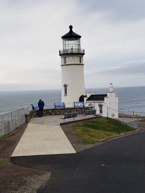 A lighthouse stands by the ocean, with a person viewing information panels nearby on a cloudy day.