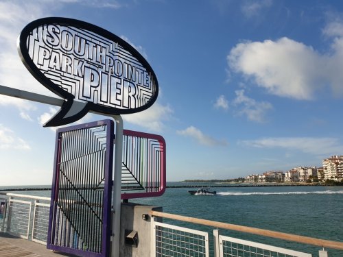 Sign for South Pointe Park Pier with a boat in the water and buildings in the background under a blue sky.