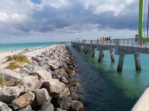 A scenic view of a pier extending over turquoise water, with people walking and rocky shoreline under a cloudy sky.