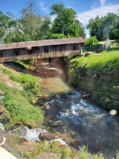 A covered bridge spans a flowing river, surrounded by lush greenery and rocky banks under a clear blue sky.