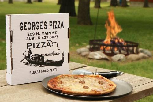 A pizza box from George's Pizza next to a plate of pizza, with a campfire in the background on a grassy area.