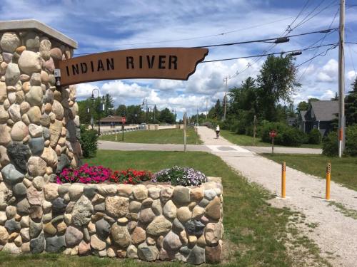 Sign reading "Indian River" at a stone entrance, with flowers and a pathway leading into a scenic area.