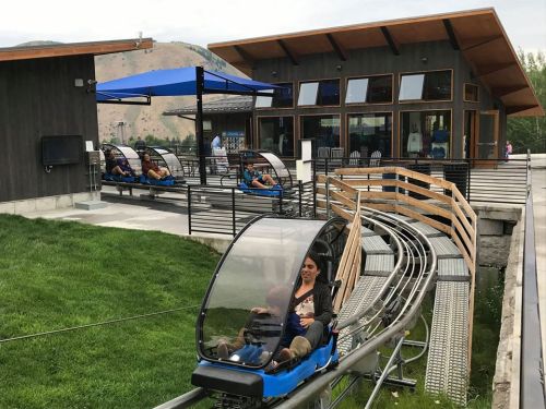 A woman and child ride a mountain coaster, with a building and grassy area in the background.