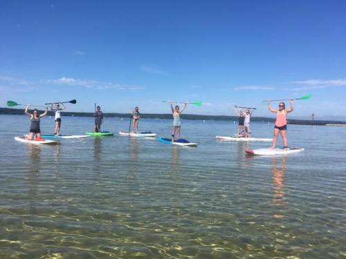 A group of people on paddleboards in calm water, holding paddles up against a clear blue sky.