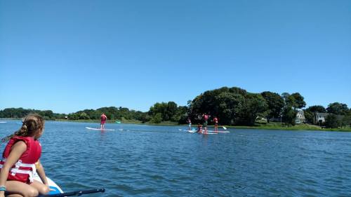 A girl in a life jacket watches a group paddleboarding on a calm lake under a clear blue sky.