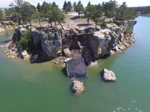 Aerial view of a rocky island with trees, surrounded by water, showing erosion and large boulders.