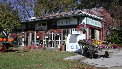 A quaint market with a brick facade, featuring flowers, pumpkins, and vintage signs, set in a rural landscape.