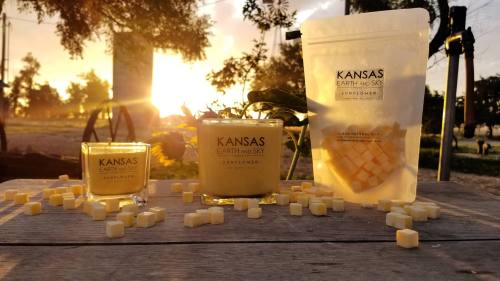 Three candles and a bag of wax melts on a wooden table, with a sunset and trees in the background.