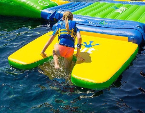A child in a life jacket climbs onto a colorful inflatable water platform from the water.