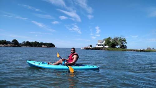 A person in a blue kayak paddles on calm water with a house and trees in the background under a clear blue sky.