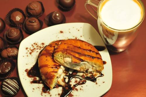 A plate with two chocolate-drizzled pastries, surrounded by assorted chocolates and a cup of frothy coffee.
