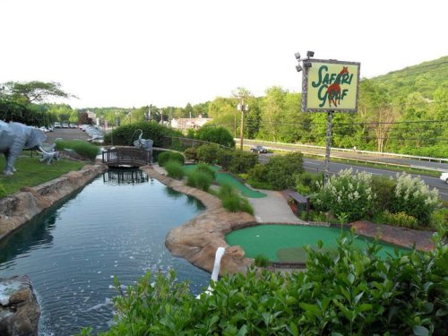 A mini-golf course with a pond, greenery, and a sign for "Safari Golf" near a road and hills in the background.