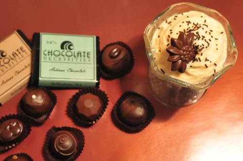 A dessert topped with chocolate and a flower, surrounded by assorted chocolates on a shiny surface.