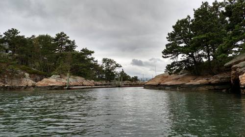 A calm waterway surrounded by rocky shores and trees under a cloudy sky.
