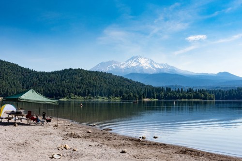 A serene lake scene with a green tent, sandy shore, and a snow-capped mountain in the background under a clear blue sky.