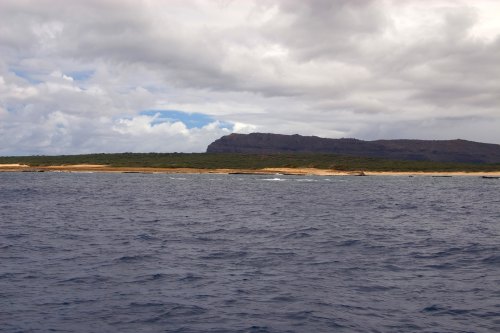 A coastal view featuring a rocky shoreline, gentle waves, and a cloudy sky over a distant mountain.