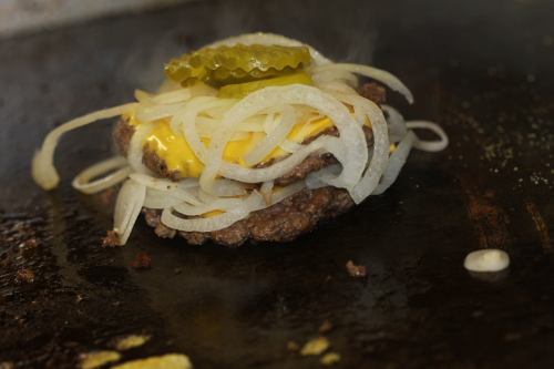 A sizzling burger with melted cheese, grilled onions, and pickles on a hot griddle.