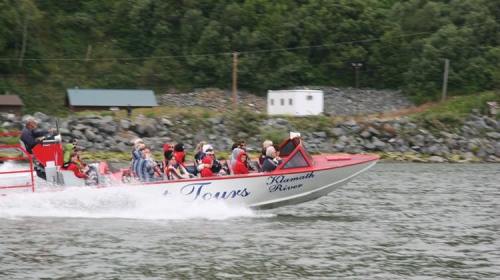 A speedboat with passengers is racing on the Klamath River, surrounded by green hills and rocky terrain.