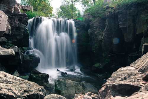 A serene waterfall cascades over rocks, surrounded by lush greenery and natural stone formations.