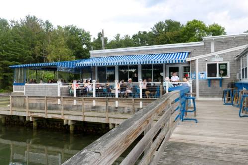 A waterfront restaurant with blue and white striped awnings, featuring outdoor seating and patrons enjoying their meals.