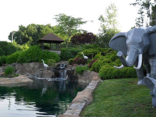A serene garden scene featuring a gray elephant statue, a pond, a waterfall, and white herons among lush greenery.
