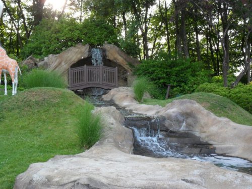 A small wooden bridge over a stream with a waterfall, surrounded by greenery and a rocky landscape.