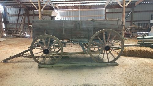A rustic wooden wagon with large wheels, displayed in a barn-like setting with various tools and materials in the background.