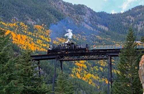 A steam locomotive crosses a trestle bridge surrounded by vibrant autumn foliage and mountains.
