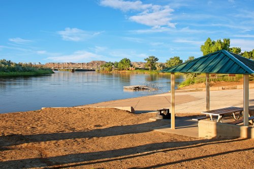 A serene river scene with a sandy shore, a picnic shelter, and a bridge in the background under a clear blue sky.
